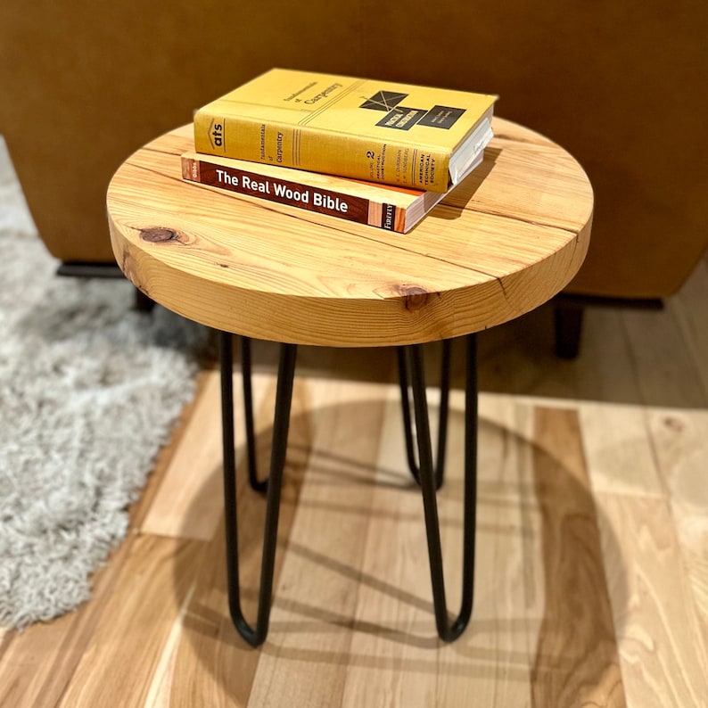 Może przedstawiać: Okrągły drewniany stolik pomocniczy z czarnymi n&oacute;żkami w kształcie szpilek. Na stole leżą dwie książki: "The Real Wood Bible" i "Fundamentals of Carpentry".