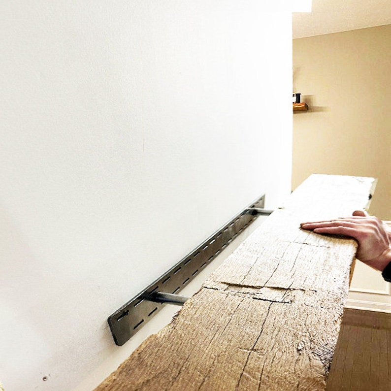 May include: A rustic wooden shelf is being installed on a white wall using metal brackets with holes for adjustable placement. The shelf is made of reclaimed wood with a natural finish.