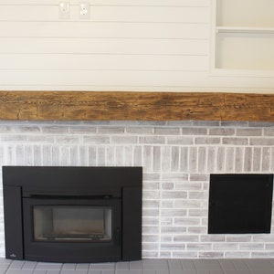 May include: A white brick fireplace with a black wood-burning stove and a wooden mantel. The fireplace is surrounded by white shiplap walls.