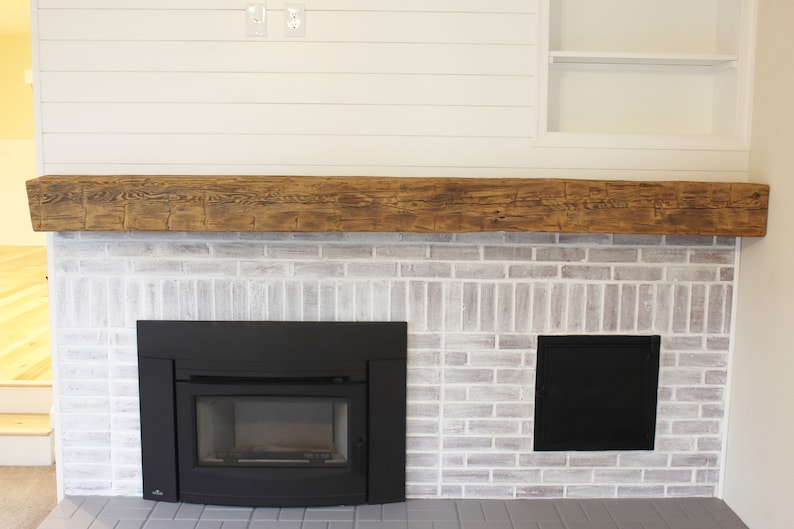 May include: A white brick fireplace with a black wood-burning stove and a wooden mantel. The fireplace is surrounded by white shiplap walls.