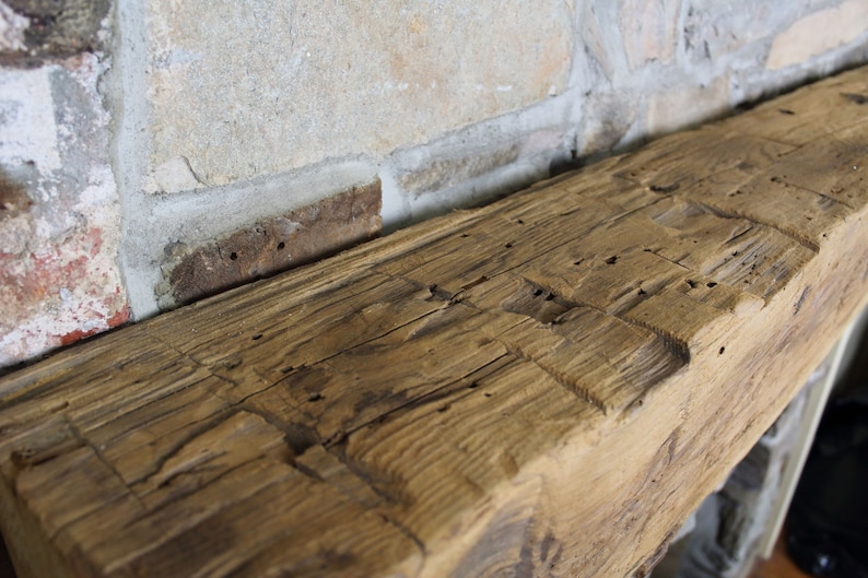 May include: Close-up of a rustic wooden mantel with a rough-hewn surface and visible knots. The wood is a warm brown color and has a distressed look.