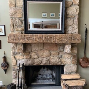 May include: A rustic wooden mantel with a black framed mirror above a stone fireplace. The fireplace has a black metal fire grate and a set of black metal fireplace tools. There are logs stacked in a metal basket to the right of the fireplace.