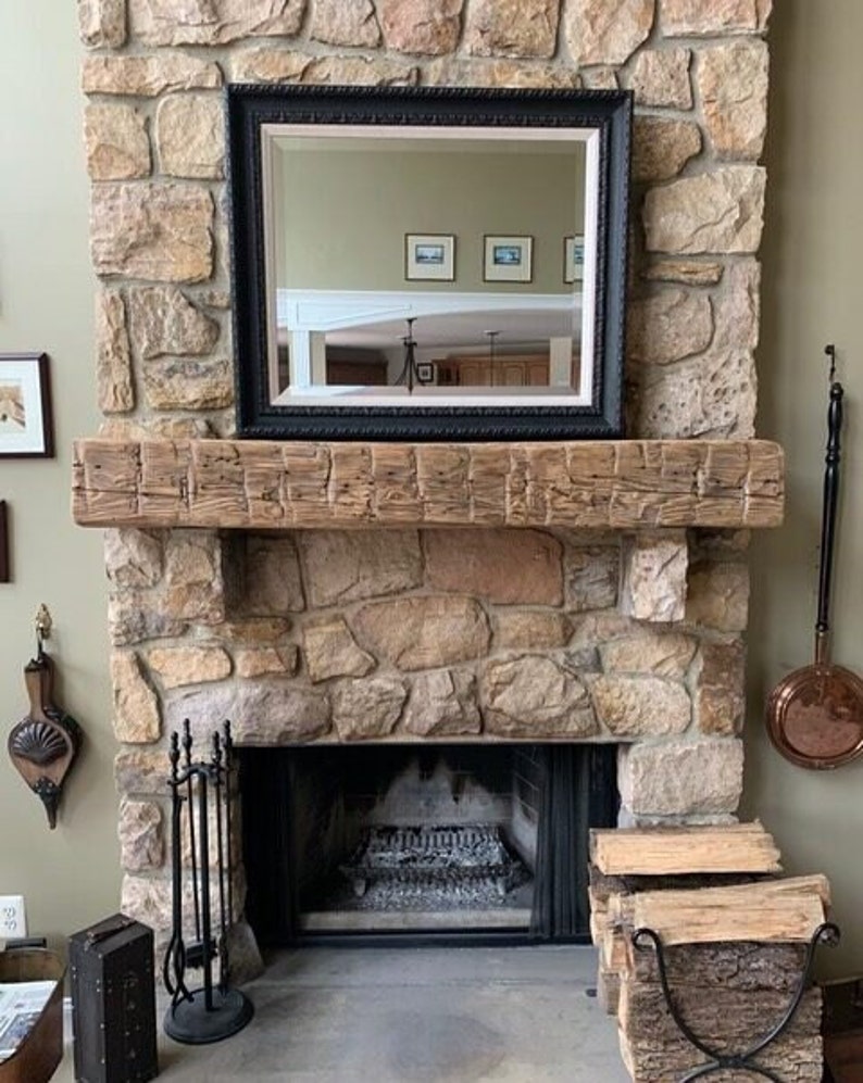 May include: A rustic wooden mantel with a black framed mirror above a stone fireplace. The fireplace has a black metal fire grate and a set of black metal fireplace tools. There are logs stacked in a metal basket to the right of the fireplace.