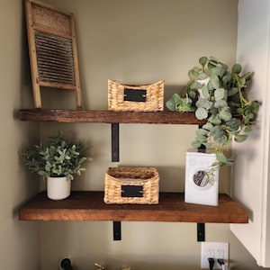 May include: Two wooden shelves with black metal brackets, mounted on a light green wall. The shelves are decorated with two wicker baskets with black labels, a small potted plant, and a vintage washboard. A white washing machine is visible below the shelves.