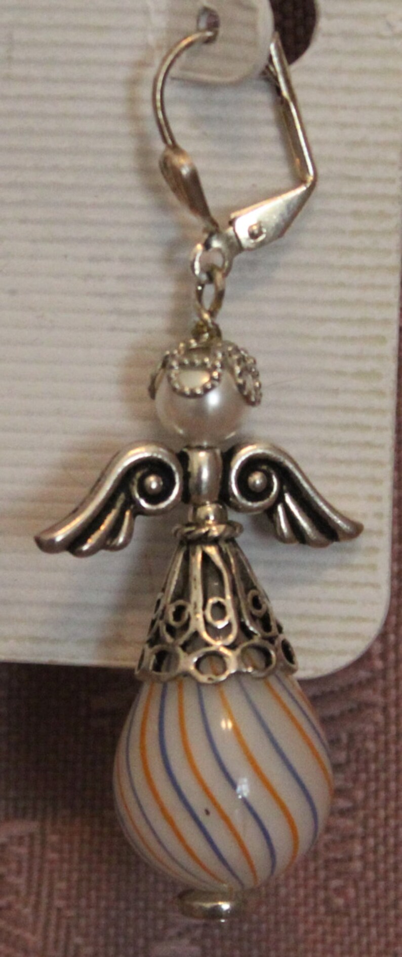 Puede incluir: Pendiente de &aacute;ngel plateado con cierre de palanca. El &aacute;ngel tiene una cabeza de perla, alas detalladas y una falda decorativa. El cuerpo en forma de l&aacute;grima es blanco con rayas naranjas y azules.