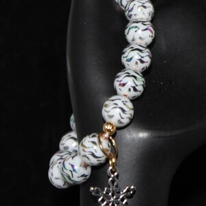 May include: White beaded bracelet with a silver snowflake charm. The beads have a zebra-like pattern in black and iridescent colors. The bracelet is held on a black display stand, showcasing the jewelry's design.