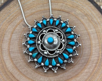 May include: A silver pendant necklace with a circular design. The pendant features a central turquoise stone surrounded by a detailed silver pattern and teardrop-shaped turquoise stones. The necklace has a silver chain.