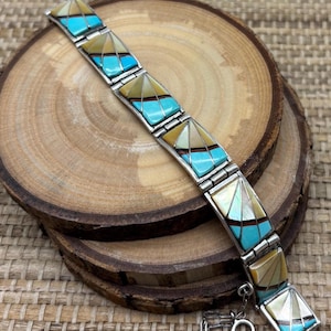 May include: A silver bracelet with turquoise, mother-of-pearl, and tan-colored triangular inlays. The bracelet is composed of rectangular links, each featuring a central turquoise section and a geometric design. The bracelet is displayed on a wooden surface.
