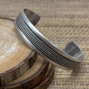May include: A silver cuff bracelet with a textured, woven design in the center. The bracelet has a simple, open design and is made of a shiny, metallic material. The bracelet is displayed on a wooden surface.