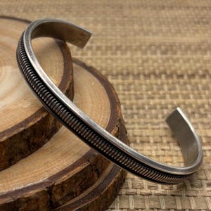 May include: A silver-toned metal cuff bracelet featuring a central raised band with a fine, vertical ribbed texture. The bracelet has a smooth, polished finish along the edges and an open design, allowing it to be slipped onto the wrist.