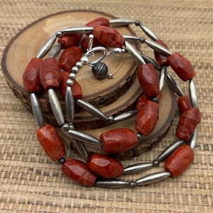 May include: A two-strand necklace with red coral beads and silver metal beads. The necklace has a silver clasp and a silver bead with a dark gray stone.