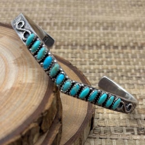 May include: A silver cuff bracelet featuring a row of small, oval-shaped turquoise stones set in a channel. The metal band displays a twisted rope texture along the edges and includes a 925 hallmark stamp near the open end of the piece.
