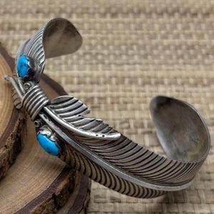 May include: A silver-toned cuff bracelet with a feather design and two turquoise-colored stones. The feather has detailed etched lines, and the bracelet has an open design. The bracelet is resting on a wooden surface.