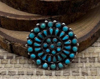 May include: A round, silver-toned pendant with a spiral design of turquoise stones. The pendant features a detailed, textured metalwork border and a central cluster of smaller turquoise stones. The turquoise stones are a vibrant blue-green color.