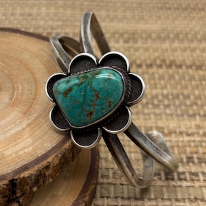 May include: A silver cuff bracelet featuring a large, heart-shaped turquoise stone set in a floral design. The turquoise stone has a mottled, blue-green color with brown veining. The bracelet has a vintage, rustic appearance.