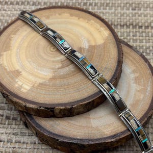 May include: A silver bracelet with inlaid rectangular segments. The segments feature a mosaic of brown, black, beige, and turquoise stones. The bracelet is displayed on a wooden surface with a natural wood grain.