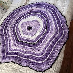 May include: A round, crocheted blanket in shades of purple, white, and black. The blanket features concentric circles of color, with a dark center and a scalloped black edge. The handmade blanket is displayed on a white bedspread.