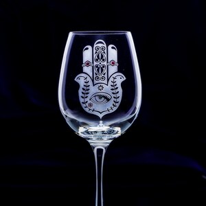 May include: A clear wine glass with a long stem, featuring an etched Hamsa hand design. The design includes an eye, floral elements, and red accents. The glass is set against a black background.