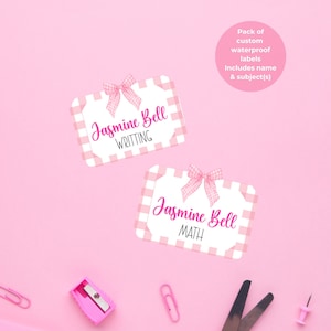 May include: Pink stationery set with custom waterproof labels. The labels feature a pink gingham pattern, a bow, and the name "Jasmine Bell" with subject titles "Writing" and "Math". Includes pink scissors, paperclips, a pencil sharpener, and push pins.
