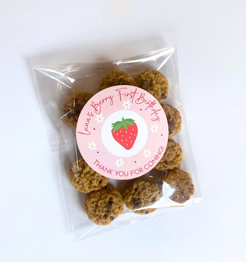K&ouml;nnte beinhalten: Ein klarer Zellophanbeutel gef&uuml;llt mit Schokoladen-Chip-Cookies. Ein runder Aufkleber auf dem Beutel tr&auml;gt die Aufschrift "Luna's Berry First Birthday" mit einer Erdbeerillustration und den Worten "Thank You For Coming!"