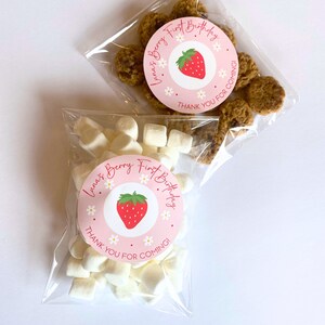 K&ouml;nnte beinhalten: Zwei transparente Zellophanbeutel gef&uuml;llt mit Leckereien. Ein Beutel enth&auml;lt Marshmallows, der andere Kekse. Jeder Beutel hat einen rosa runden Aufkleber mit einer Erdbeerillustration und dem Text "Luna's Berry First Birthday, Thank You For Coming!"