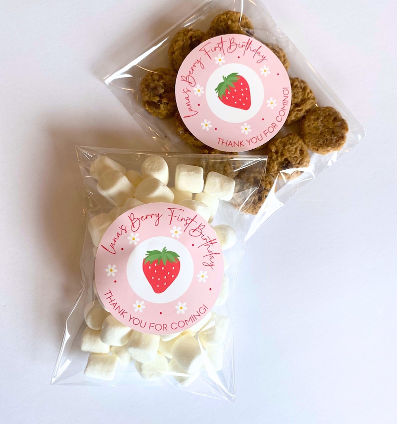 K&ouml;nnte beinhalten: Zwei transparente Zellophanbeutel gef&uuml;llt mit Leckereien. Ein Beutel enth&auml;lt Marshmallows, der andere Kekse. Jeder Beutel hat einen rosa runden Aufkleber mit einer Erdbeerillustration und dem Text "Luna's Berry First Birthday, Thank You For Coming!"