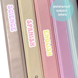 May include: A close-up of several pastel-colored binders with labels. The binders are labeled "SCIENCE", "SPANISH", and "ENGLISH". A blue circle with the text "Waterproof subject labels" is also visible.