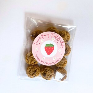 K&ouml;nnte beinhalten: Ein klarer Cellophanbeutel gef&uuml;llt mit kleinen, runden Keksen. Ein rosa Aufkleber mit einer Erdbeerillustration und dem Text "Luna's Berry First Birthday" ist auf der Vorderseite. Der Text "Thank you for coming!" ist ebenfalls vorhanden.