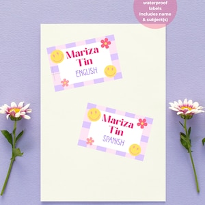 May include: Two custom waterproof labels with the name "Mariza Tin" and subject labels in English and Spanish. The labels have a pink and purple checkered border, smiley face and flower graphics. The text "Pack of custom waterproof labels" is also visible.