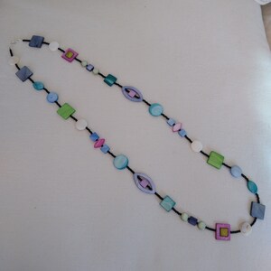 Op de afbeelding: Een lange ketting met een zwart koord en kleurrijke, geometrische kralen. De kralen zijn in tinten blauw, groen, roze en wit.