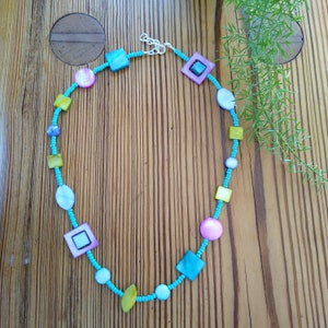 Op de afbeelding: Een turquoise kralenketting met verschillende gekleurde en gevormde schelpkraaltjes, waaronder vierkanten, cirkels en ovalen. De ketting ligt op een houten oppervlak.