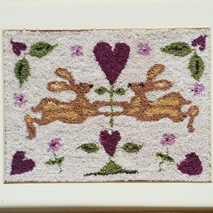 May include: A framed needlepoint art piece featuring two golden-brown rabbits facing each other, centered between two purple hearts. The design includes small green leaves and purple flowers on a textured off-white background, encased in a gold frame.