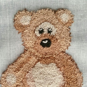 Op de afbeelding: Een bruine pluche teddybeer met witte accenten op de buik, poten en oren. De beer zit op een lichtgrijze stoffen achtergrond.