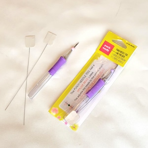 May include: A purple and silver punch needle tool with a spacer bead. The packaging includes the text "Punch Needle Tool and Threader" and "L'Aguille de Poncon et L'Enfile-Aiguille".