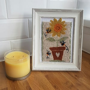 May include: A white framed picture featuring a needlepoint design of a sunflower in a brown pot with a heart cut-out. Three black and orange bees are flying around the sunflower.