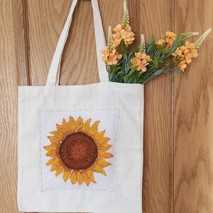 May include: A white canvas tote bag featuring a square patch with a detailed embroidered sunflower in shades of yellow and brown. The patch is attached with visible orange stitching. A bouquet of orange artificial flowers sits inside the bag.