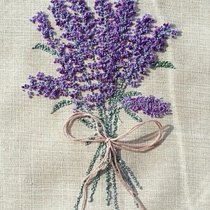 Könnte beinhalten: Nahaufnahme einer handgestickten Stickerei eines Straußes Lavendelblüten, die mit einem braunen Band auf einem beigefarbenen Leinenstoff gebunden sind.