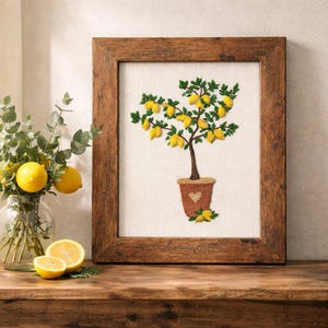 May include: A framed embroidery of a lemon tree in a brown pot with a heart design. The artwork is in a wooden frame and sits on a wooden table. A vase of lemons and greenery is to the left.