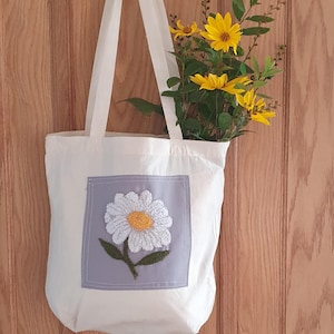 Op de afbeelding: Een witte canvas tote bag met een grijze vierkante patch met een geborduurde witte madeliefje met een gele kern en groene bladeren.