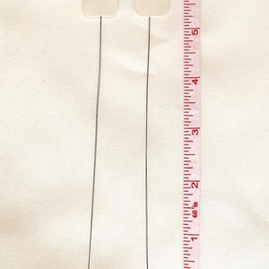 May include: Two white square tags with black wire stems, measuring approximately 6 inches in length, are displayed against a white background with a ruler for scale.