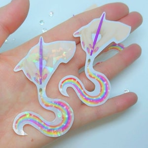May include: Two iridescent holographic stickers of a hand painting a rainbow with a paintbrush. The stickers are on a white background.