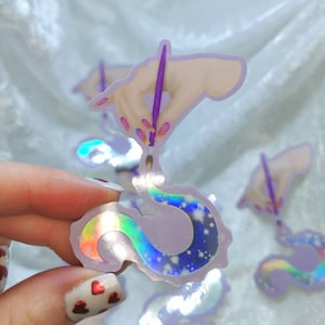 May include: Holographic sticker showing a hand with a purple paintbrush, creating a swirling design with a starry night sky and iridescent details. The sticker has a pale purple border and is held by a person.