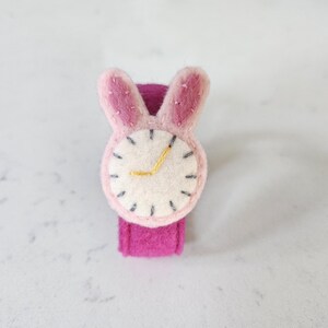 Puede incluir: Un reloj de pulsera de fieltro rosa con una esfera blanca y orejas de conejo. La esfera tiene costuras negras alrededor del borde y una manecilla de hora dorada.