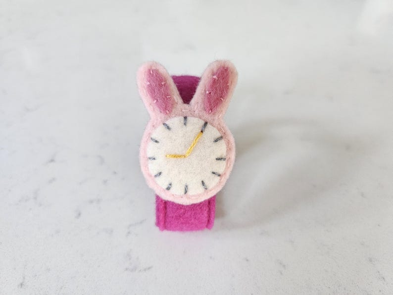 Puede incluir: Un reloj de pulsera de fieltro rosa con una esfera blanca y orejas de conejo. La esfera tiene costuras negras alrededor del borde y una manecilla de hora dorada.