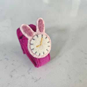 Puede incluir: Un reloj de pulsera de fieltro rosa con una esfera blanca y orejas de conejo. La esfera tiene marcas negras y las manecillas son doradas.
