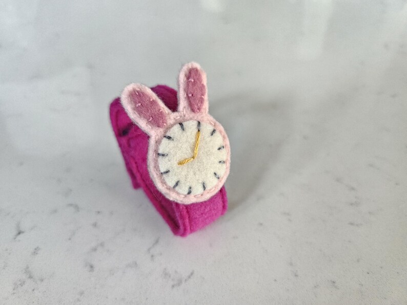 Puede incluir: Un reloj de pulsera de fieltro rosa con una esfera blanca y orejas de conejo. La esfera tiene marcas negras y las manecillas son doradas.