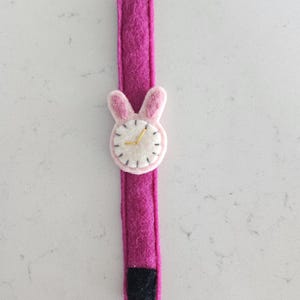 Puede incluir: Un reloj de pulsera de fieltro rosa con una cara de conejo de fieltro blanco y agujas de reloj doradas. El reloj tiene un cierre de velcro negro.