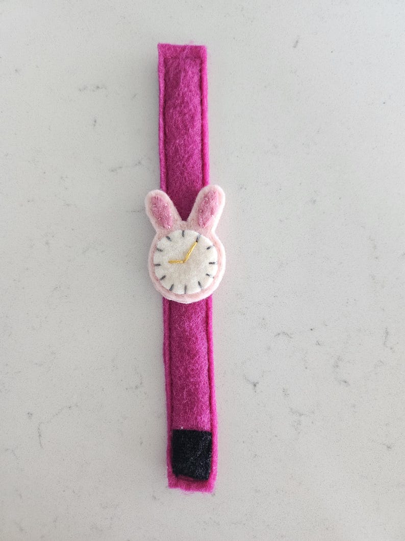 Puede incluir: Un reloj de pulsera de fieltro rosa con una cara de conejo de fieltro blanco y agujas de reloj doradas. El reloj tiene un cierre de velcro negro.