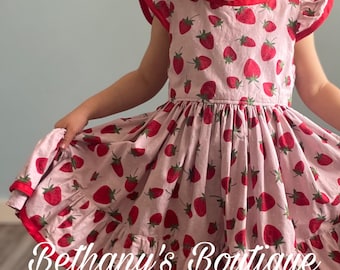 May include: A light pink dress with a red strawberry pattern and red trim. The dress has short sleeves with red trim and a ruffled hem. The text "Bethany's Boutique" is printed on the bottom of the dress.
