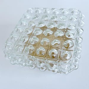 May include: A square clear glass light fixture cover featuring a grid of raised, rounded bubble-like protrusions on its surface. The glass has a thick, textured appearance with a subtle golden-brown tint visible through the centre of the piece.